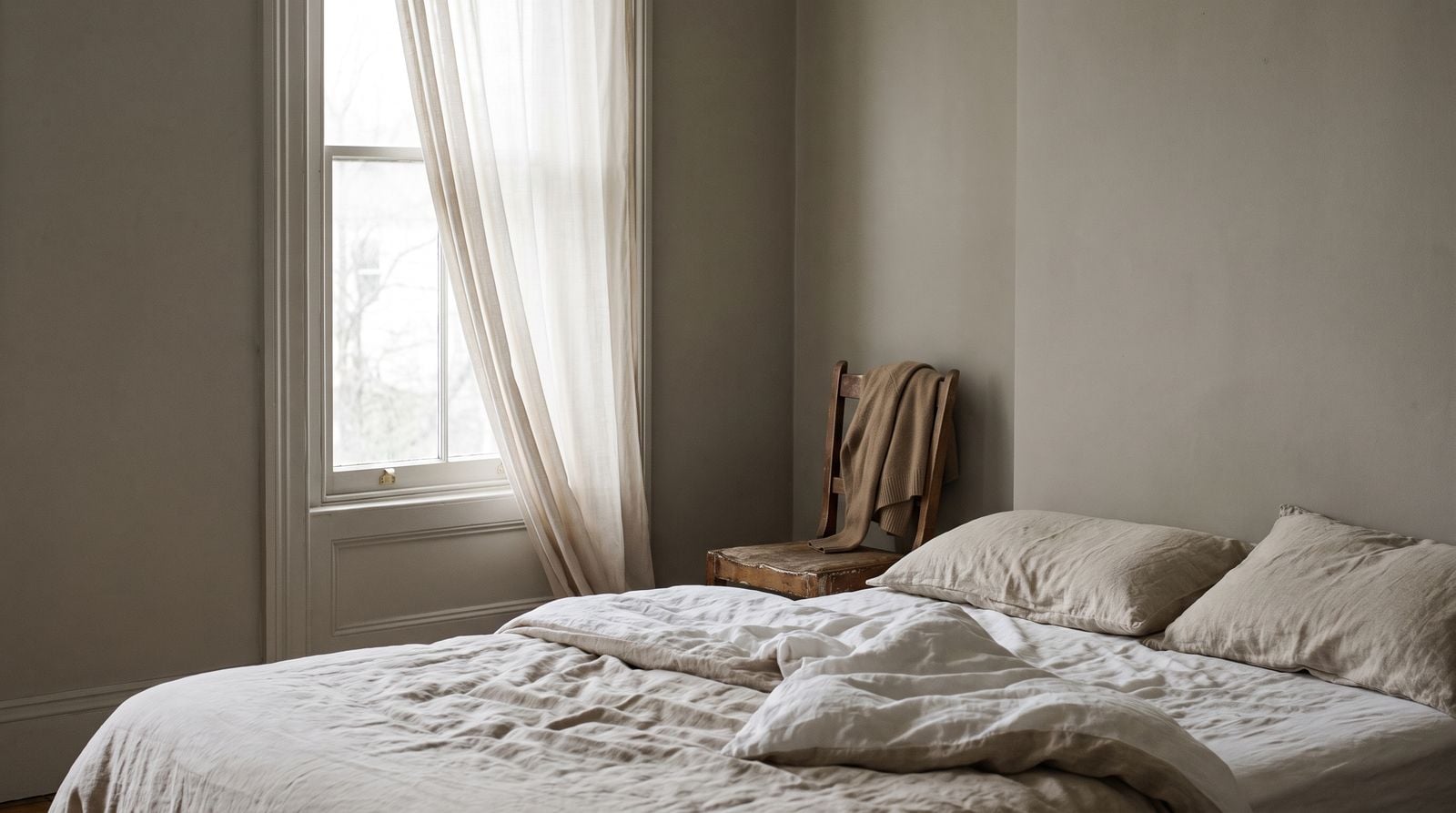 An unmade bed in a plain room, daylight coming through a tall window.